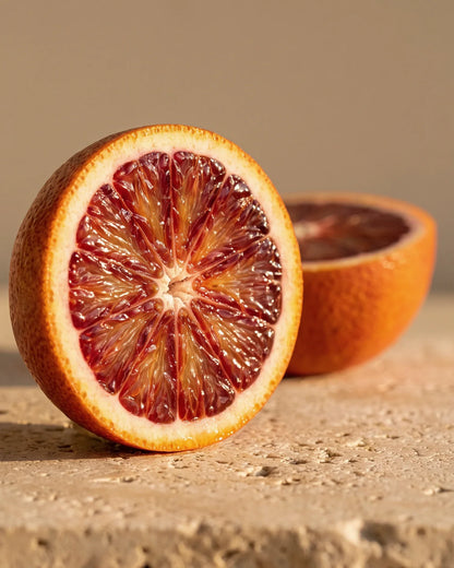 A halved blood orange with deep ruby flesh on warm travertine — the sweet, berry-like citrus scent of Delune Blood Orange Pure Essential Oil