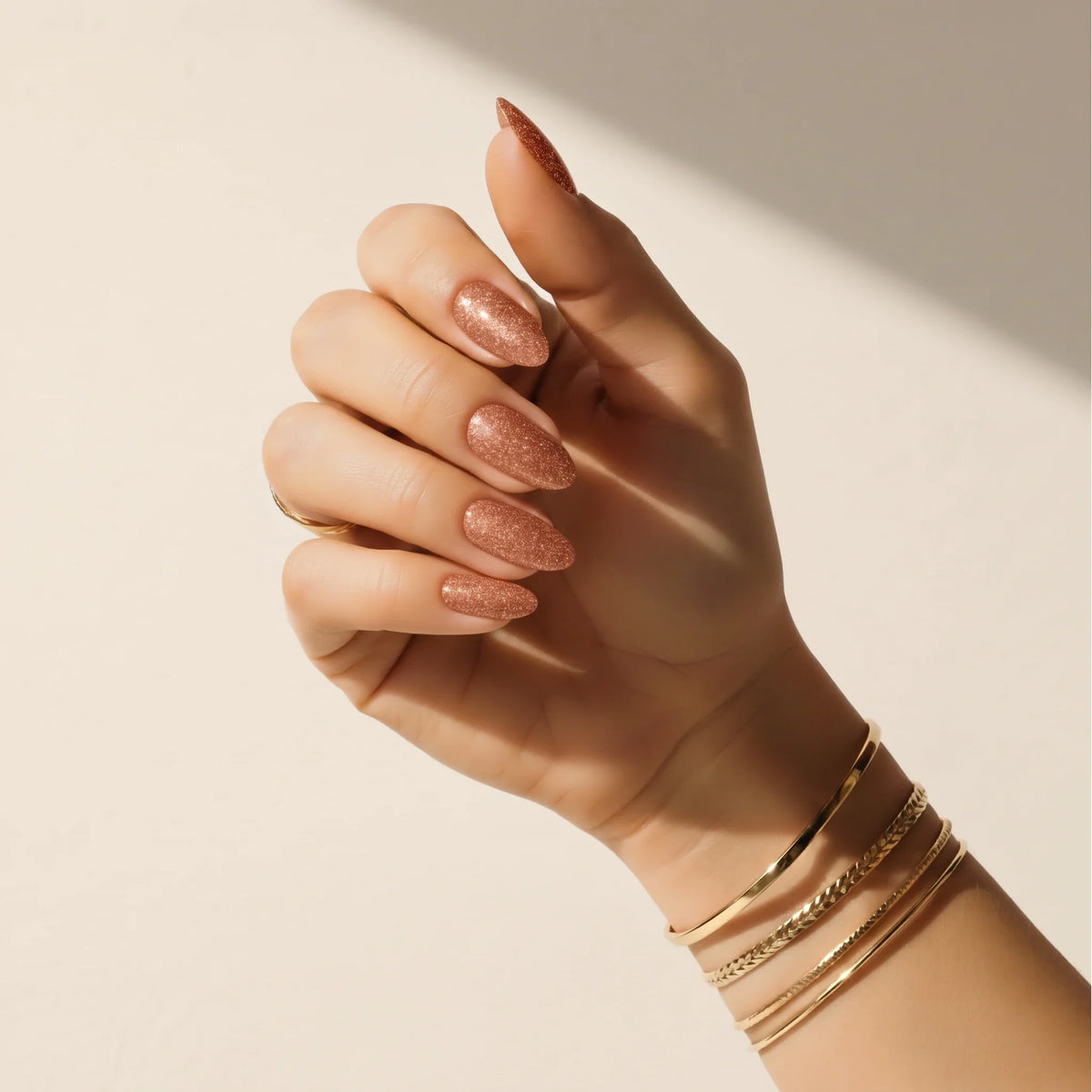 Hand wearing Delune Copper Glitter nail wraps with gold bangles against a warm plaster wall