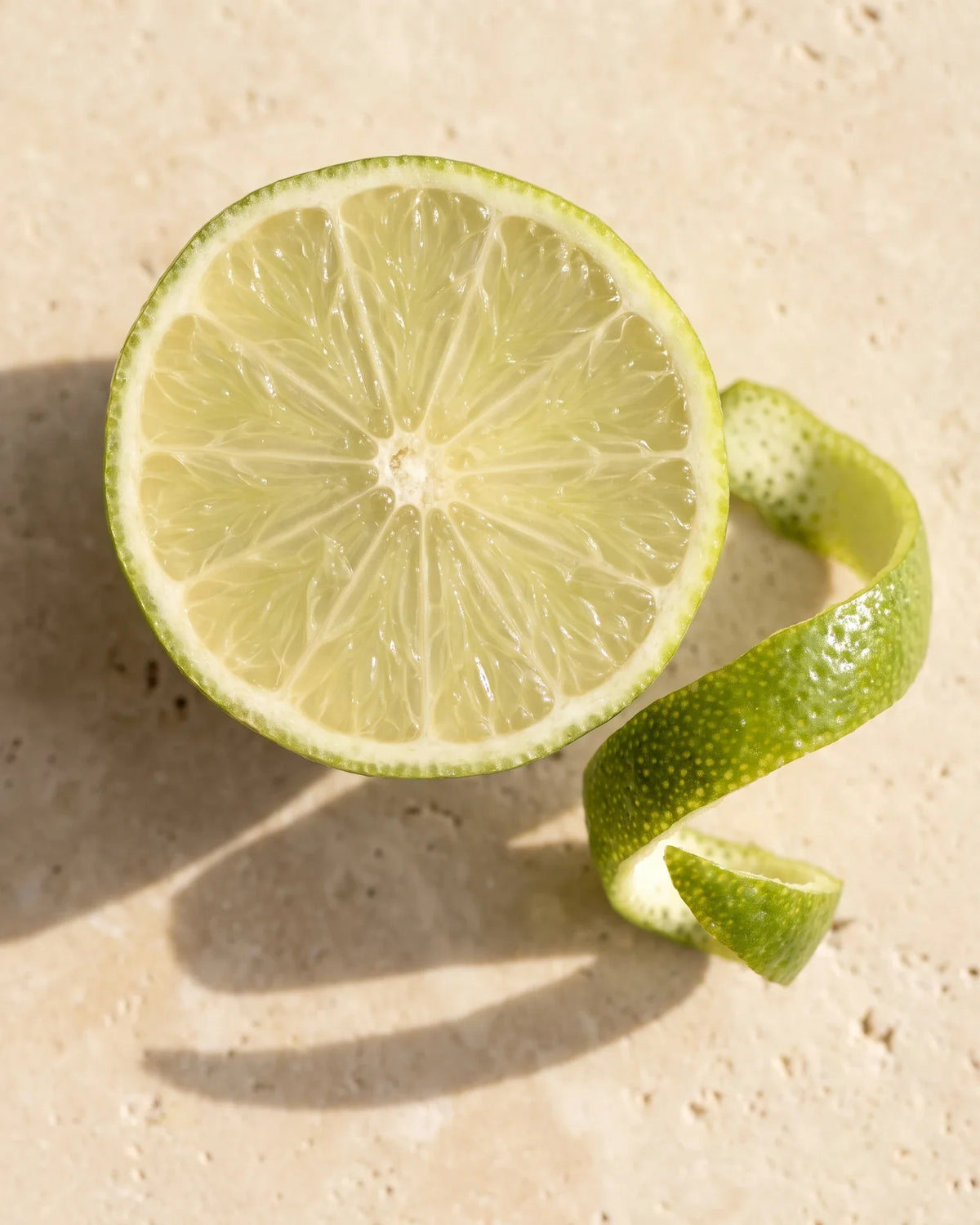 A halved fresh lime and a curl of lime zest seen from above on warm travertine — the sharp, zesty scent of Delune Lime Pure Essential Oil